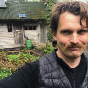 Oliver Broadbent standing smiling in front of the Longhouse, a timber building at Hazel Hill Wood to illustrate the post 'in praise of the longhouse'