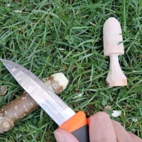 a hand in the frame holds a specialised whittling knife to a stick, with a whittled mushroom nearby