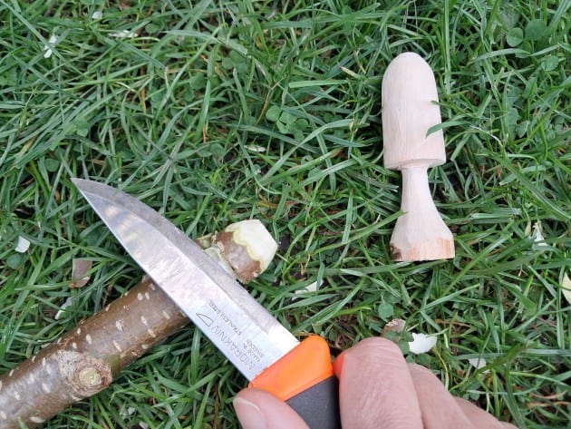 a hand in the frame holds a specialised whittling knife to a stick, with a whittled mushroom nearby